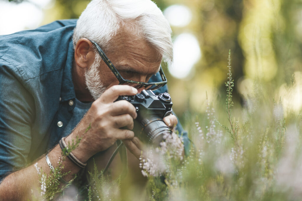 A keen gray-haired man photographs flowers in the garden. Hobby grandfather on summer vacation. He is engaging in creative activities for seniors in Assisted Living at The Delaney at The Vale.
