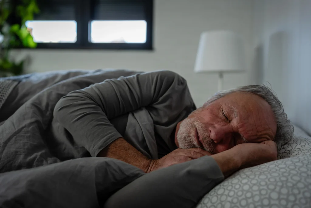 An elderly man with grey bed sheets getting 7-9 hours from his home at The Delaney at The Vale in Woburn, MA.