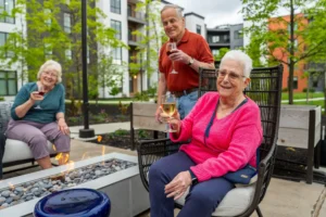 Seniors at The Delaney® at The Vale relax outdoors around a warm gas fire pit with glasses of wine, embracing an engaging community lifestyle outside the modern residential building.
