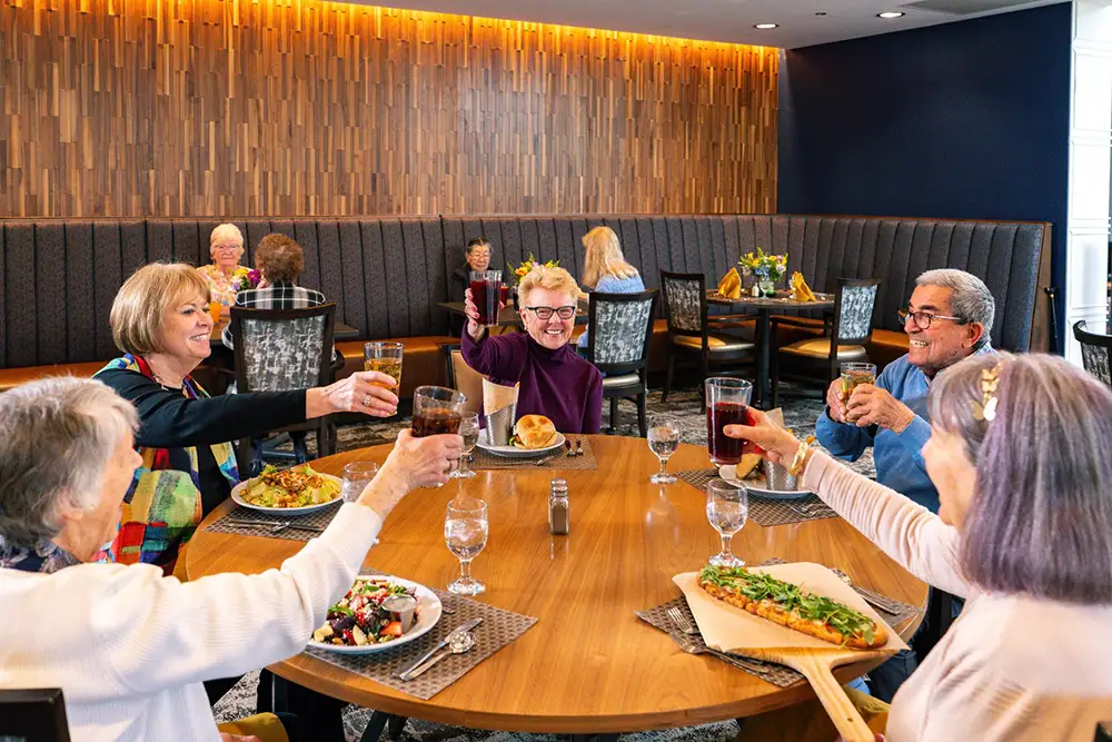 A lively group of seniors at The Delaney® at The Vale toast their drinks around a large dining table filled with food, enjoying the vibrant community lifestyle in a modern restaurant-style venue.
