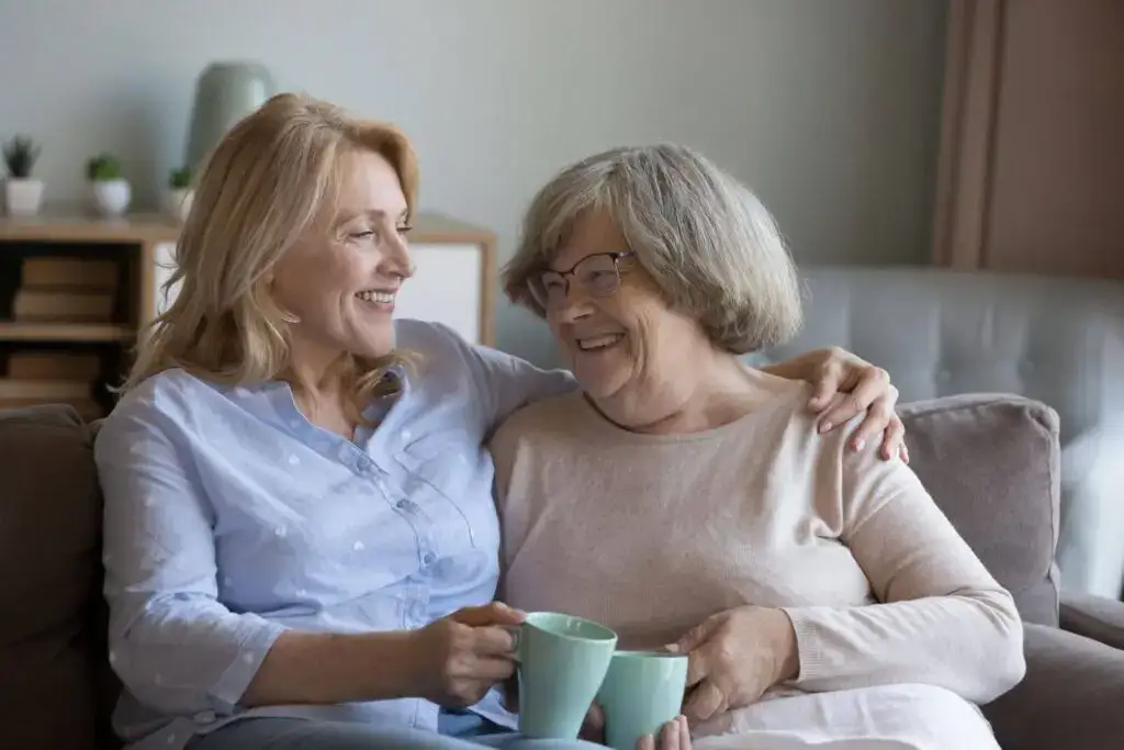 An adult daughter and her senior mother hugging, smiling, sitting on a couch while discussing common signs of caregiver burnout and a need for assisted living in Woburn, MA.